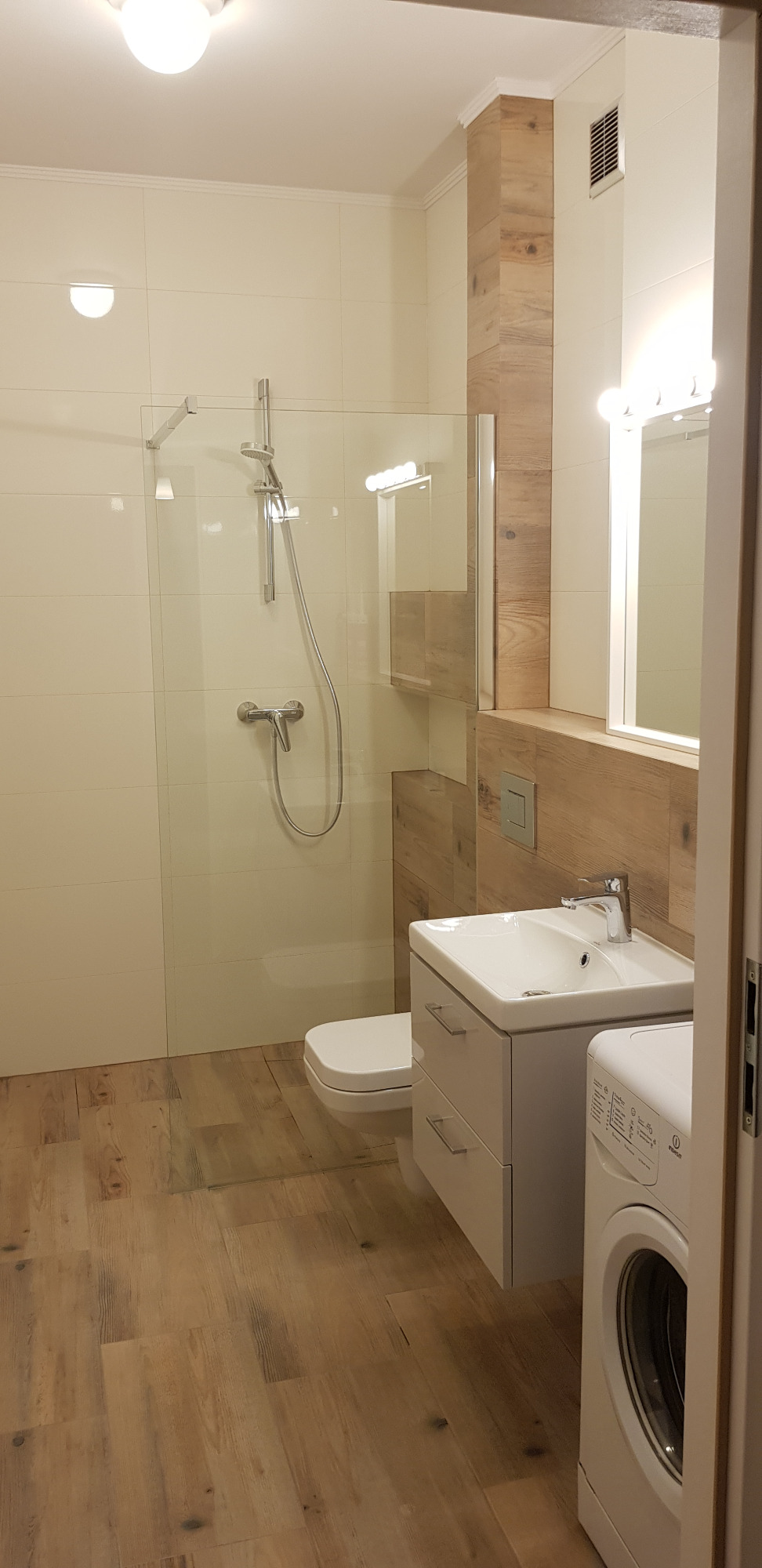 Jasna łazienka z kabiną prysznicową, umywalką z szafką, toaletą i pralką, wykończona płytkami imitującymi drewno na podłodze i częściowo na ścianach.