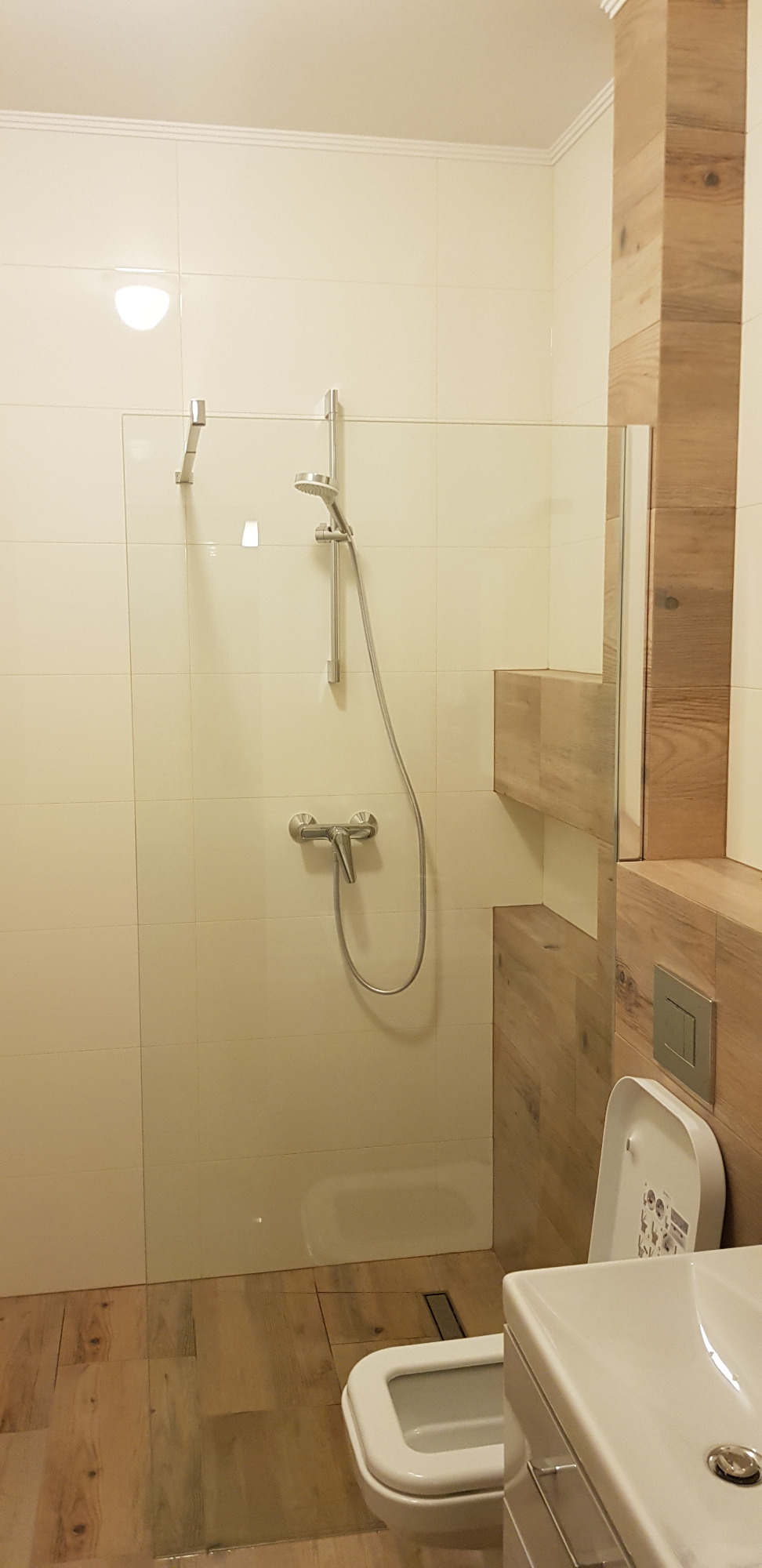 Nowoczesna łazienka z kabiną prysznicową typu walk-in, wykończona płytkami imitującymi drewno na podłodze i części ściany, armatura chromowana, widoczny fragment toalety i umywalki.