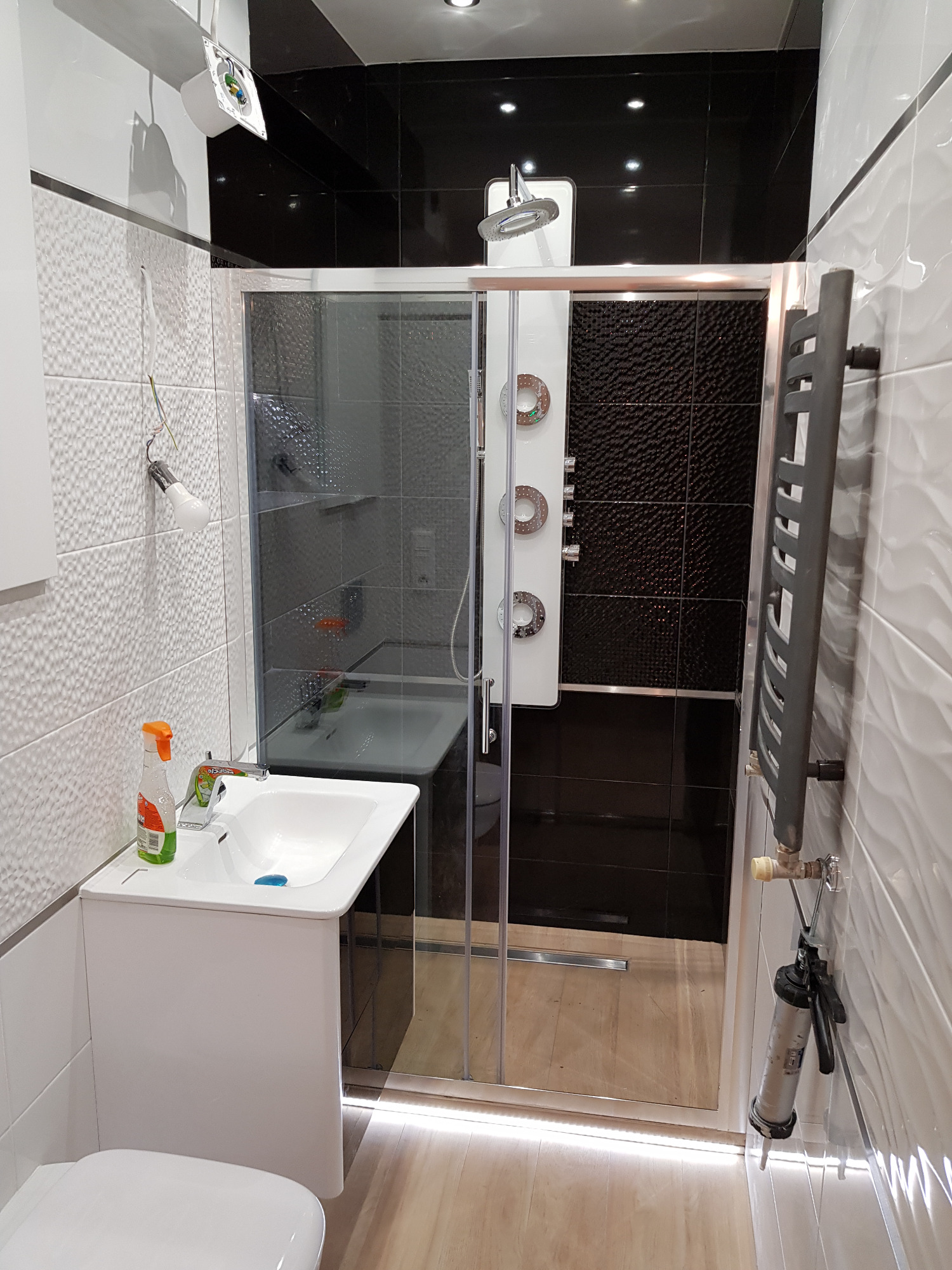 Nowoczesna łazienka z kabiną prysznicową typu walk-in, czarno-białymi płytkami, wiszącą szafką z umywalką i grzejnikiem drabinkowym; widoczny silikon w tubie.