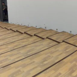 Układanie jasnych paneli podłogowych z ciemnymi obramowaniami, widoczny pracownik w tle przy drzwiach, biała ściana z gniazdkami.