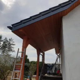 Zadaszenie tarasu z drewnianą konstrukcją nośną i nowym, ciemnym pokryciem dachowym, widoczne z poziomu gruntu, z drabiną opartą o słup.