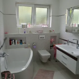 Jasna łazienka z wanną, toaletą, umywalką z szafką i oknem, wykończona białymi płytkami z dekoracyjnymi półkami z kosmetykami i kwiatami.