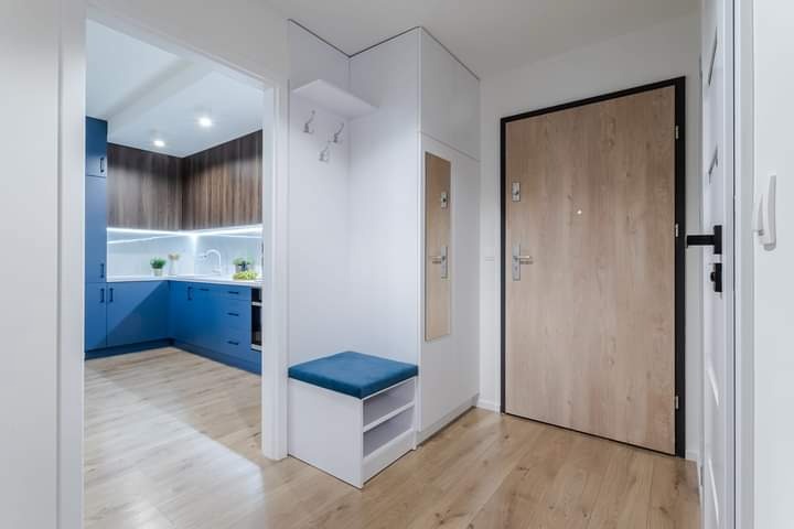 Przedpokój z szafą w zabudowie, siedziskiem z niebieską poduszką i lustrem, widok na niebieską kuchnię z drewnianymi szafkami górnymi.