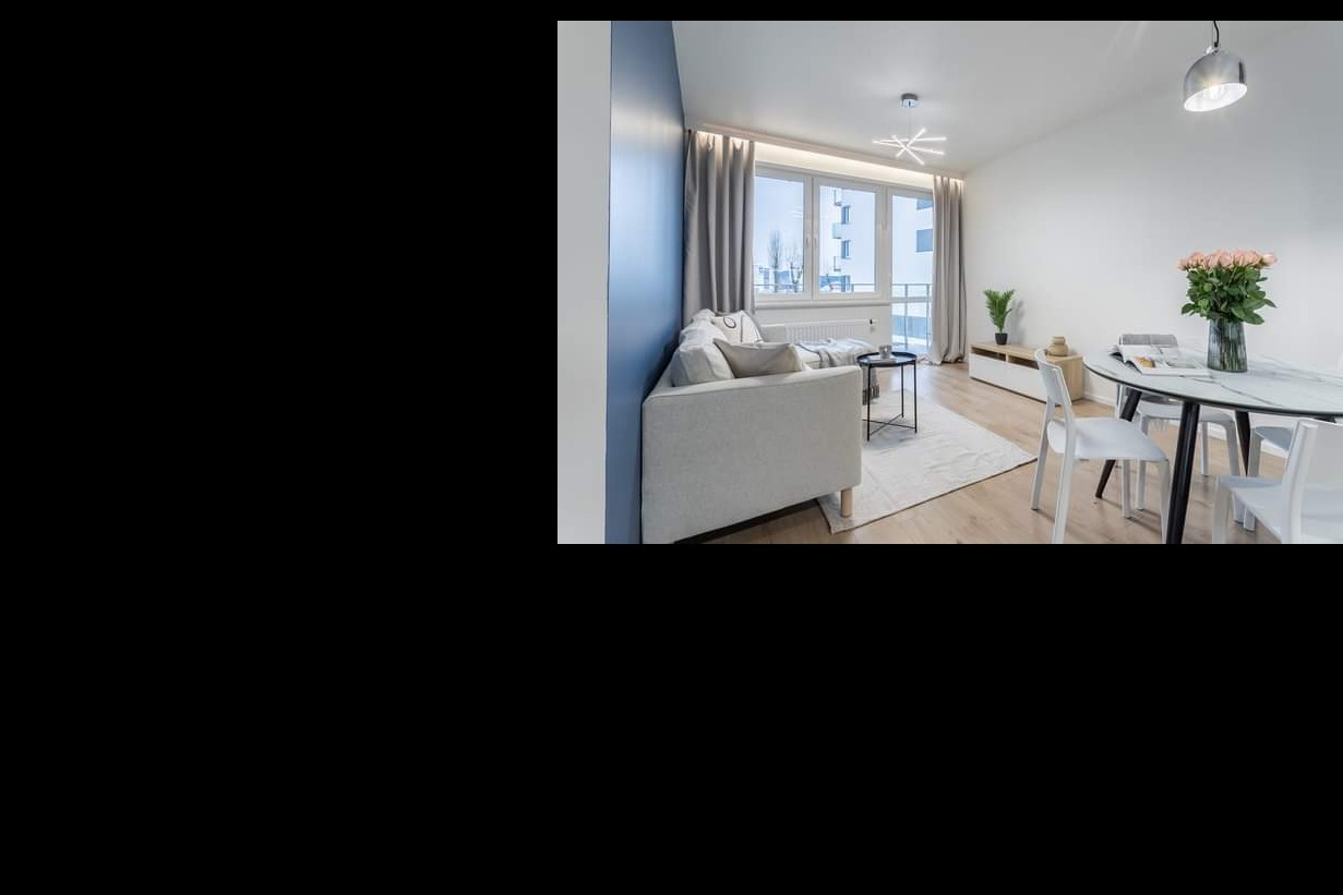 Jasne wnętrze salonu z akcentem w postaci granatowej ściany, beżowa sofa z poduszkami, okrągły stolik kawowy na dywanie, stół z marmurowym blatem i białymi krzesłami, bukiet róż w szklanym wazonie.