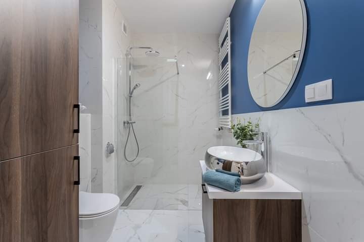 Nowoczesna łazienka z kabiną prysznicową typu walk-in, umywalką w kształcie misy i szafką z drewnopodobnym frontem, wykończona białym marmurem z szarymi żyłami i akcentami w kolorze granatowym.