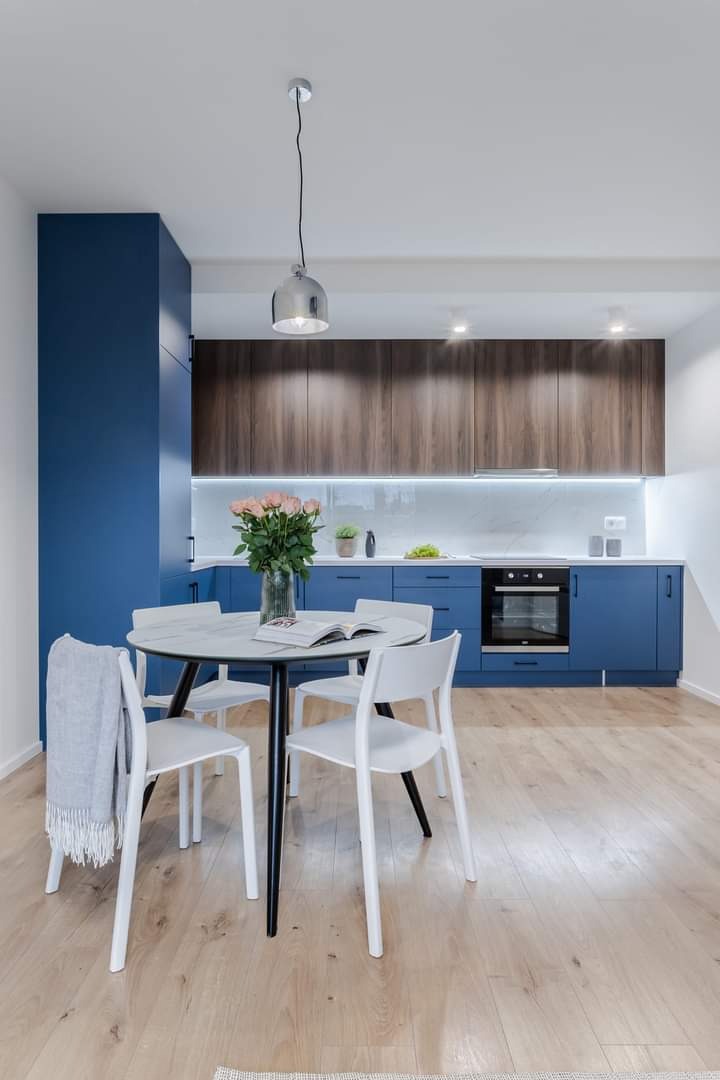 Nowoczesna kuchnia z niebieskimi szafkami, drewnianymi górnymi szafkami, marmurowym blatem i okrągłym stołem z bukietem różowych róż