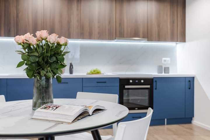 Nowoczesna kuchnia z niebieskimi szafkami dolnymi i drewnopodobnymi górnymi, blatem z białego marmuru, oświetleniem LED pod szafkami i bukietem różowych róż w szklanym wazonie na okrągłym stole...
