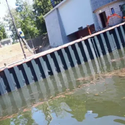 Brzeg rzeki umocniony pionowymi, pomalowanymi na ciemno szaro żelaznymi profilami, z małą pomarańczową koparką na górze, budynek w tle, odbicie nieba w wodzie.