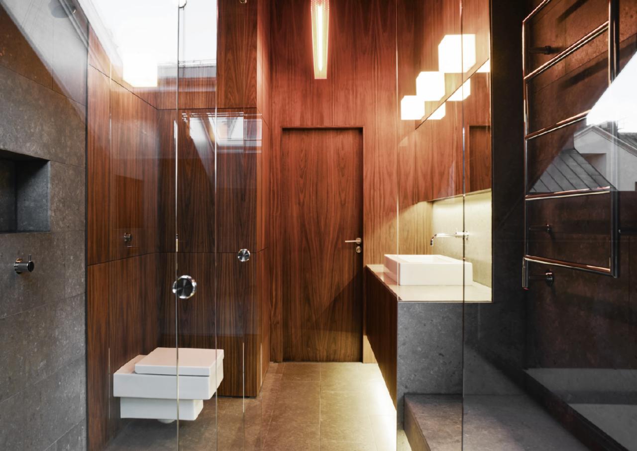 Nowoczesna łazienka z drewnianymi panelami, szklaną kabiną prysznicową i minimalistyczną białą ceramiką, oświetlona designerskimi lampami.