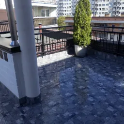 Wykończony taras na dachu z ciemnymi płytkami imitującymi kamień, otoczony czarną balustradą, z widokiem na budynki mieszkalne i tuję w donicy.