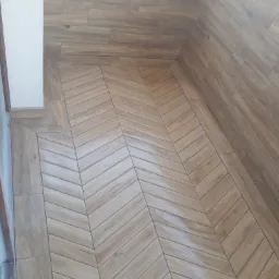 Ukośne ułożenie płytek imitujących drewno na podłodze i ścianie balkonu, widoczny narożnik i biała ściana.