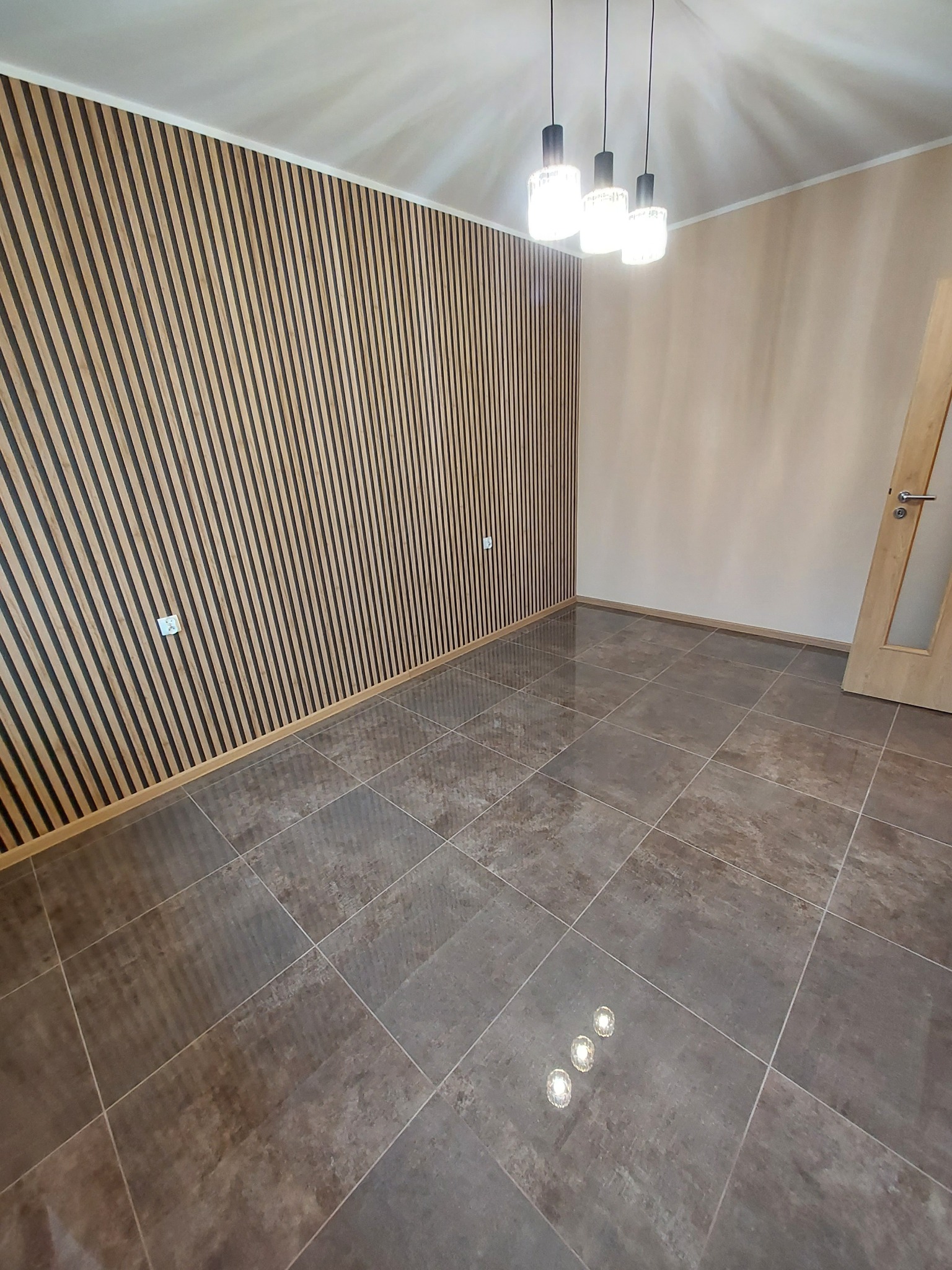 Pusta przestrzeń z brązowymi płytkami podłogowymi, drewnianą ścianą z listew i trzema lampami wiszącymi, prezentująca nowoczesne wykończenie wnętrza.