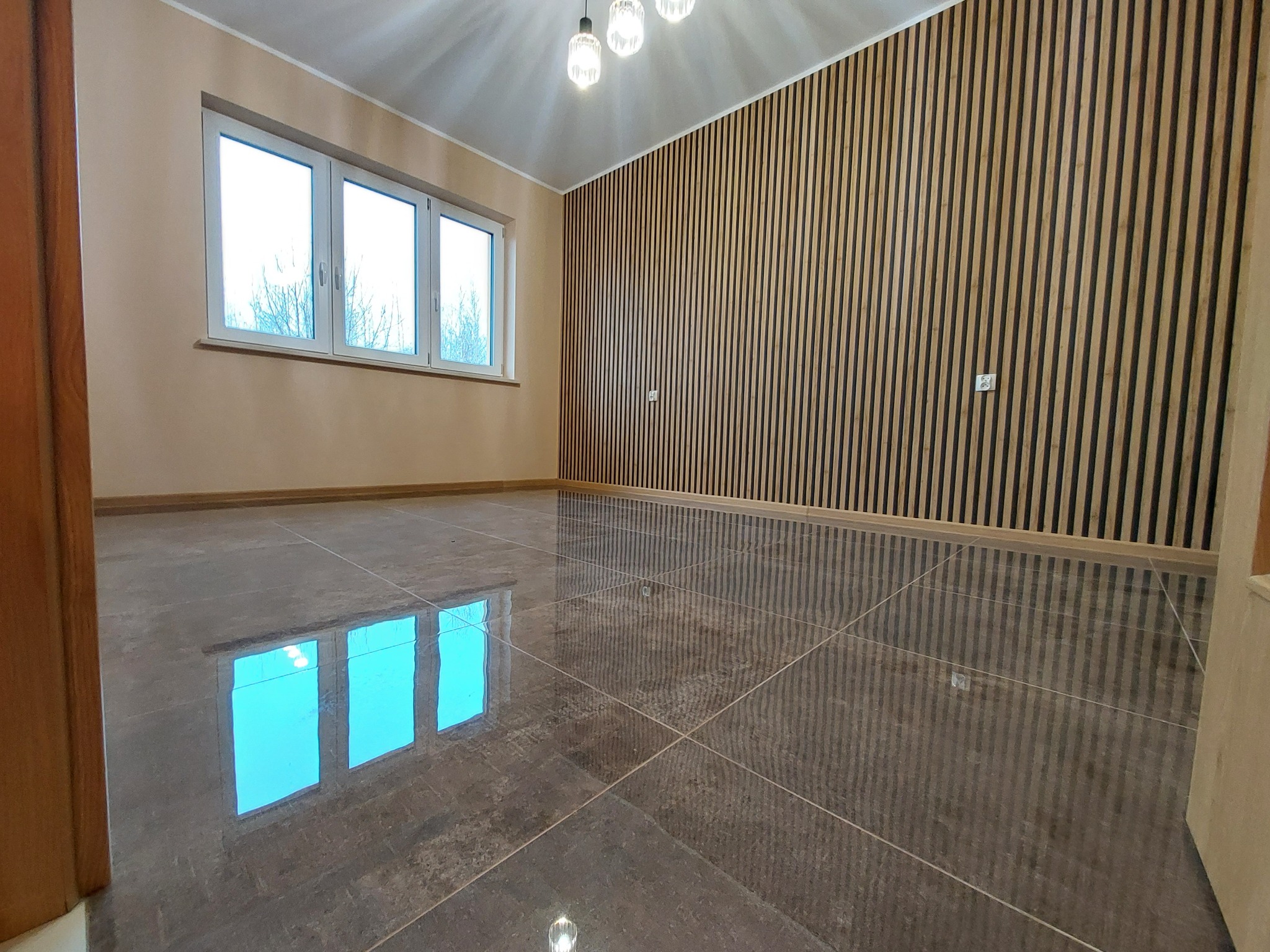 Pokój z brązowymi płytkami podłogowymi odbijającymi okno, ściana z drewnianych paneli pionowych, trzy lampy sufitowe.