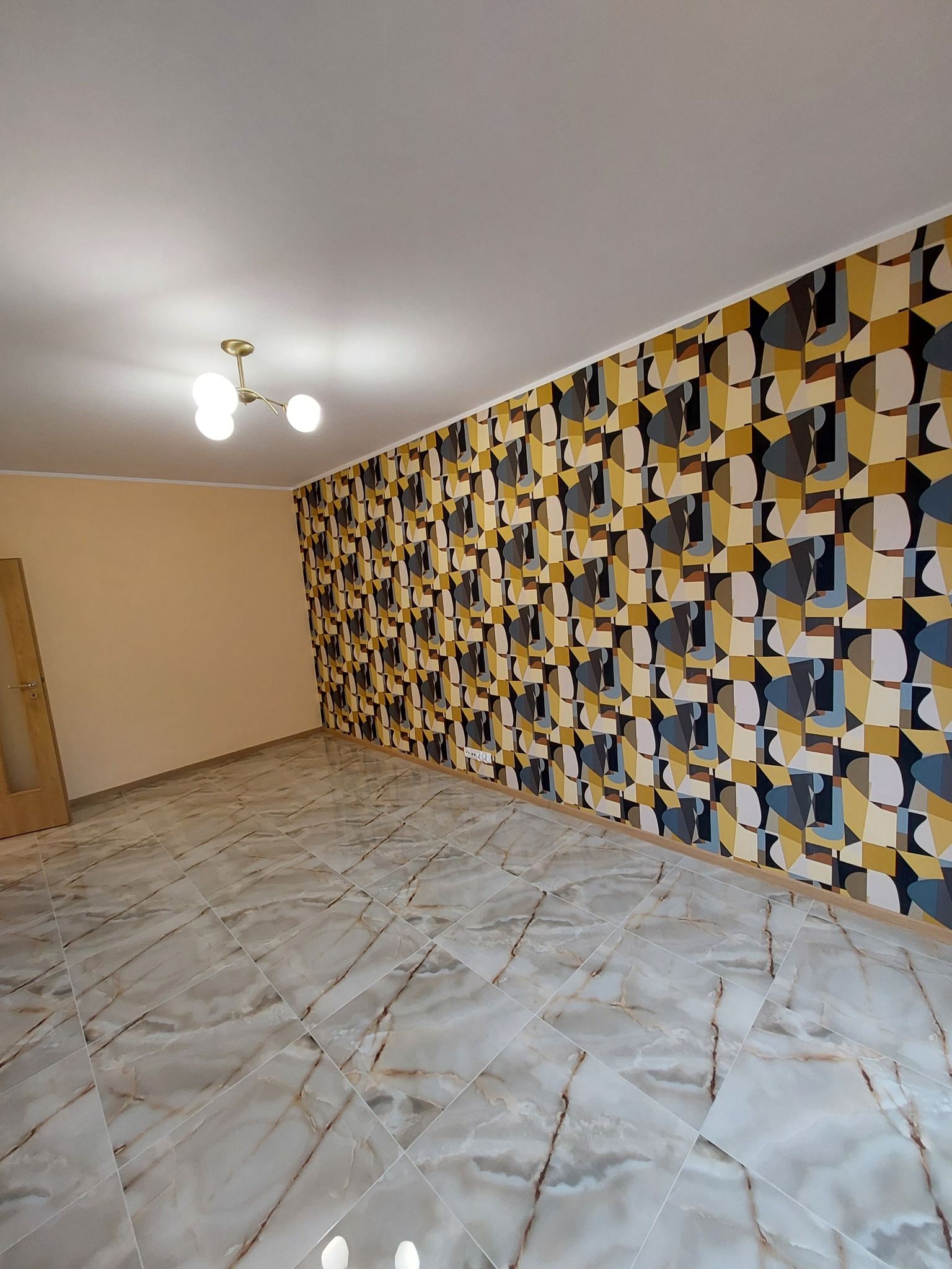 Pokój po remoncie z marmurową podłogą ułożoną w karo i ścianą z geometryczną tapetą w odcieniach żółci, szarości i czerni, oświetlony lampą sufitową z trzema kloszami.