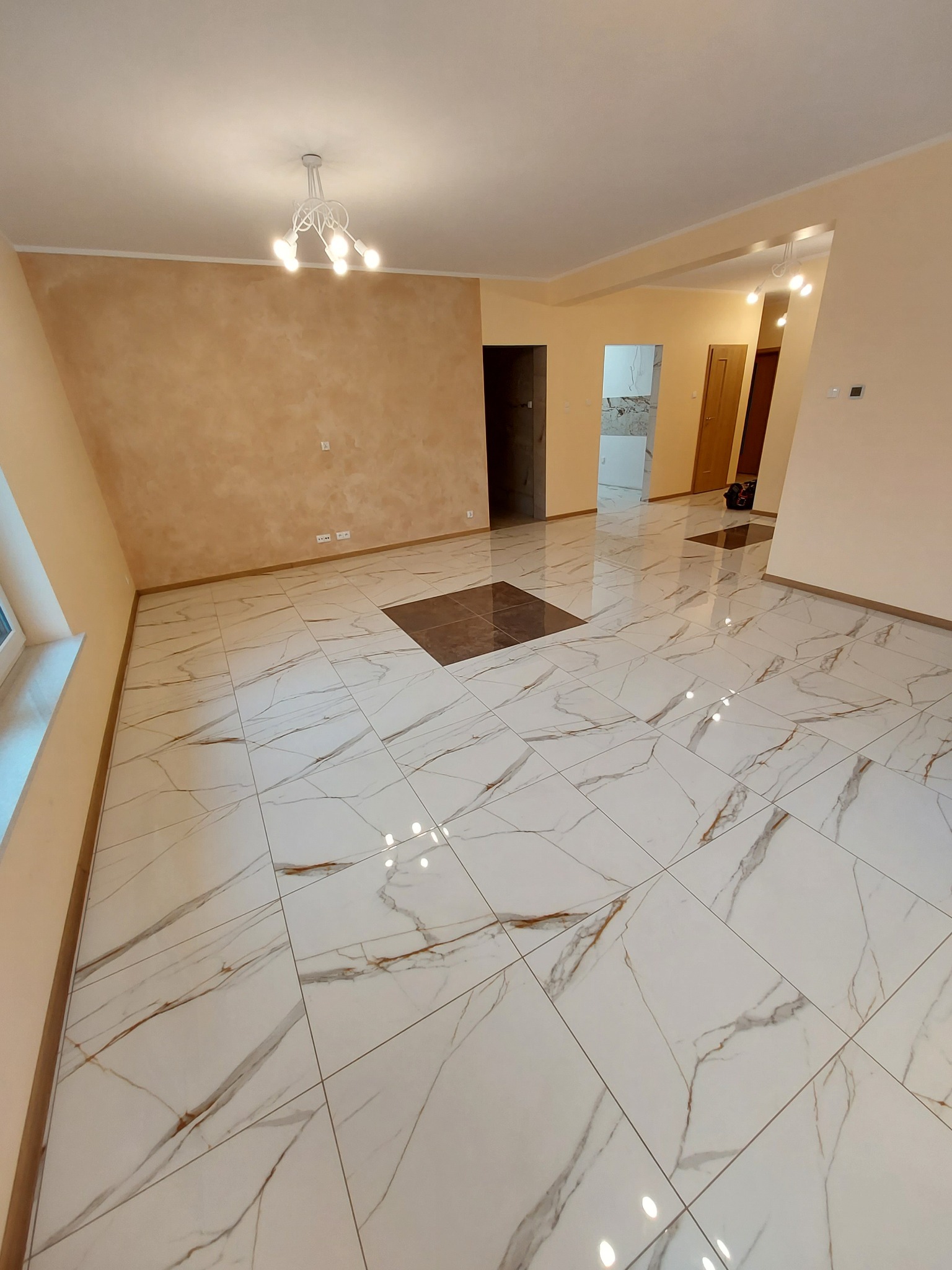 Wykończone wnętrze: przestronny salon z marmurową podłogą w odcieniach bieli i brązu, ściany w beżowej kolorystyce z dekoracyjną fakturą, widoczne drzwi do innych pomieszczeń i designerski żyrandol.