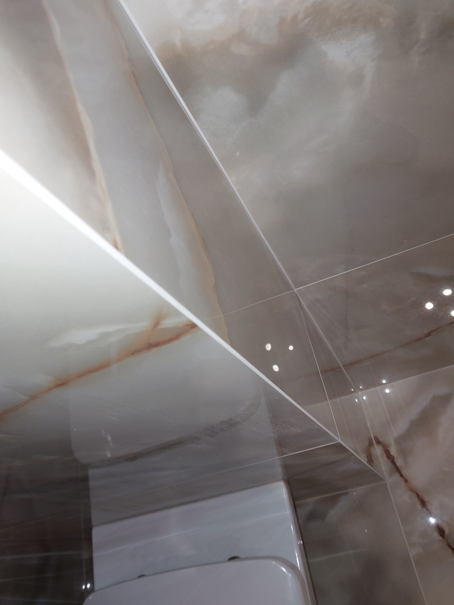 Błyszczące, beżowo-brązowe płytki imitujące marmur na ścianie łazienki, widoczny fragment toalety, oświetlenie odbijające się w powierzchni płytek.