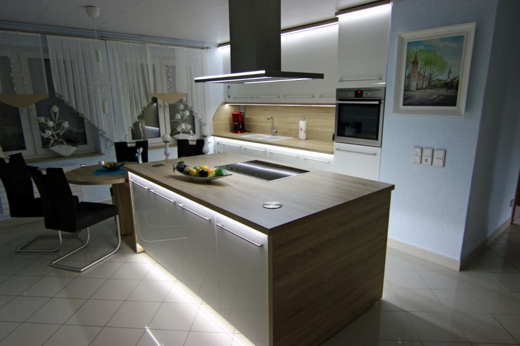 Nowoczesna kuchnia z wyspą o drewnianym blacie i białych frontach, podświetlana listwami LED, z płytą indukcyjną i owocami na blacie.