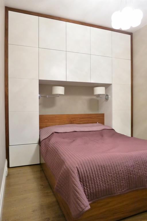 Sypialnia z łóżkiem i zabudową ścienną z białymi szafkami, drewniana rama i zagłówek, lampki nocne