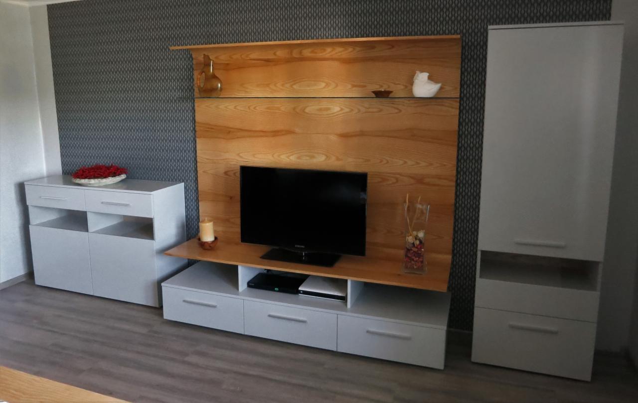 Zestaw mebli na wymiar w salonie: drewniana ściana TV z półkami i szafką RTV, biała komoda z kwiatami oraz wysoka szafa wnękowa w szarym odcieniu, na tle wzorzystej tapety.