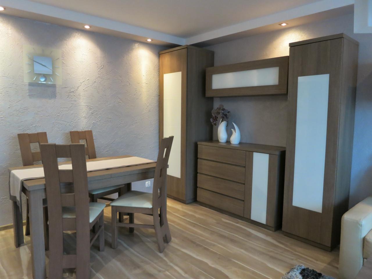 Jadalnia z drewnianym stołem i krzesłami, zabudową meblową w odcieniach brązu i beżu z białymi wstawkami, oświetleniem punktowym i dekoracyjnym zegarem na ścianie o fakturze baranka.