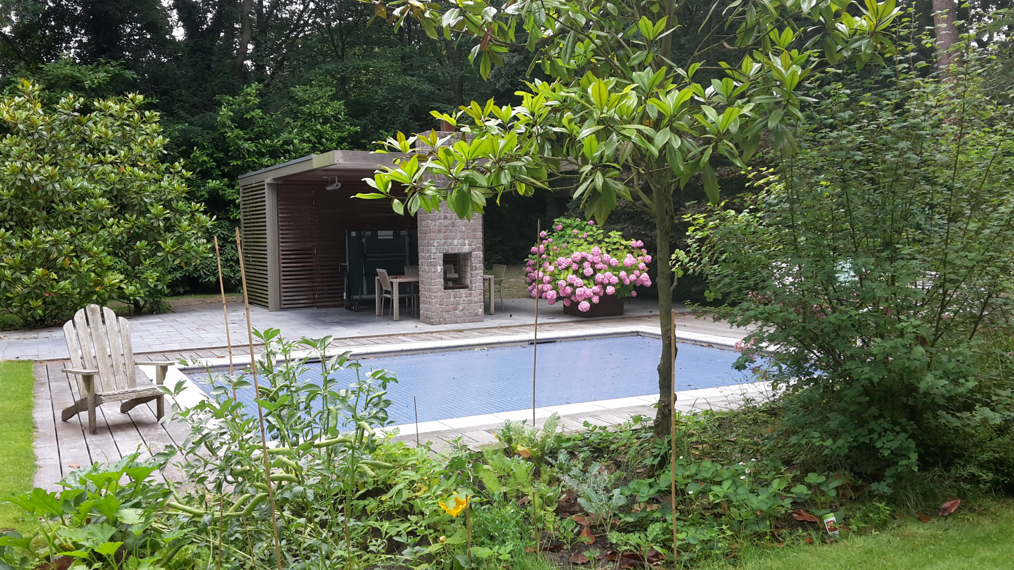 Przydomowy basen z niebieskim pokryciem, otoczony zielenią, z drewnianym krzesłem Adirondack po lewej i altaną z ceglanym kominkiem w tle.