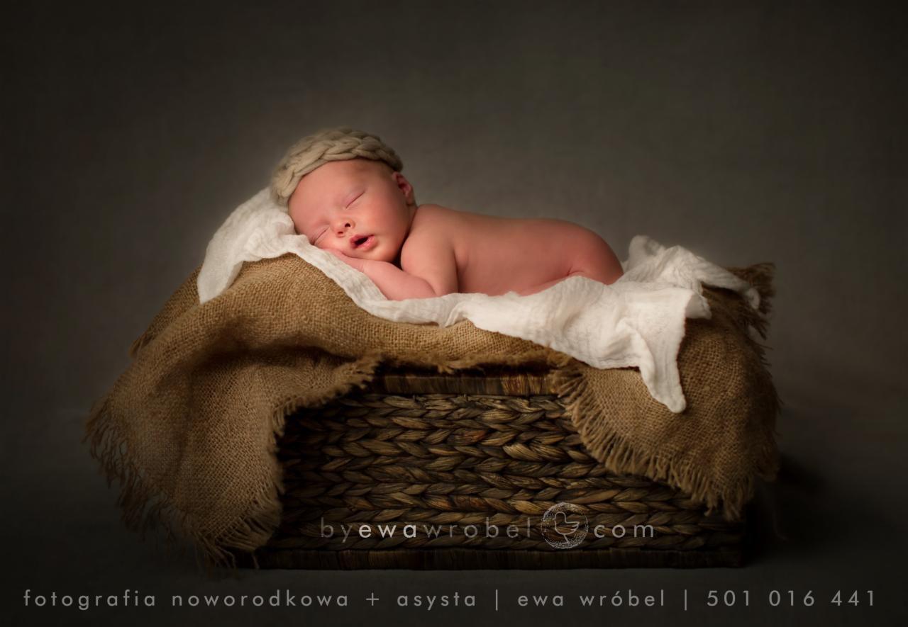 Noworodek śpiący w koszyku wyłożonym jutą i białym materiałem, na głowie ma dzierganą czapeczkę w odcieniach beżu.