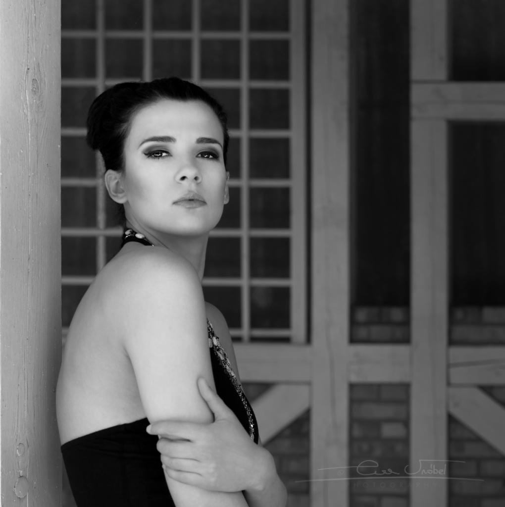 Czarno-biały portret kobiety z mocnym makijażem oczu, opierającej się o drewniany filar na tle okna z kratownicą.