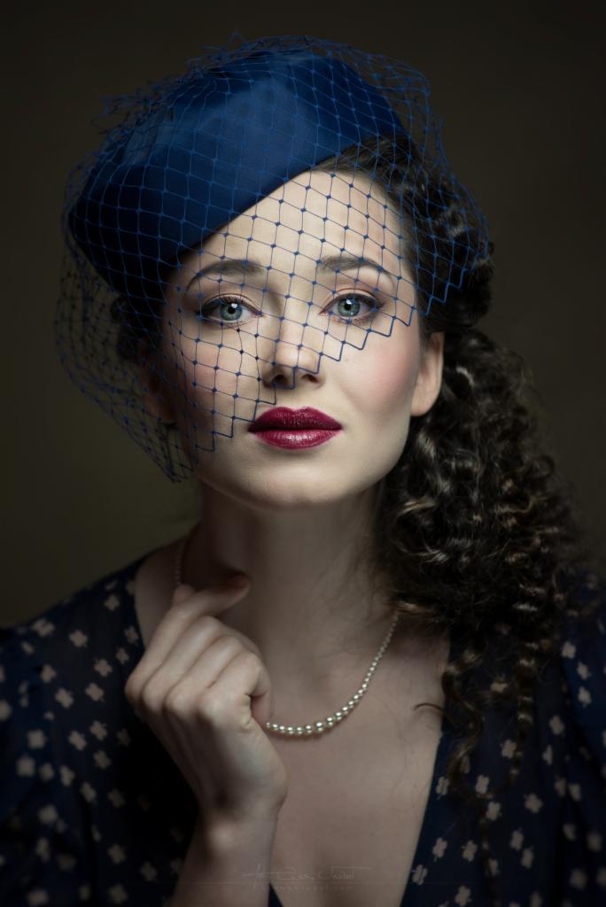Portret kobiety w niebieskim kapeluszu z woalką, perłowym naszyjnikiem i ciemnoczerwoną szminką, z delikatnym uśmiechem i dłonią przy brodzie.