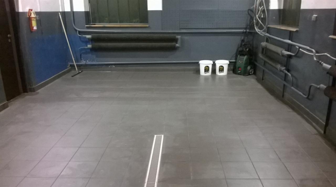 Wnętrze myjni z szarymi płytkami na podłodze, odpływem liniowym, dwoma białymi wiaderkami z grafiką niedźwiedzia, myjką ciśnieniową i gaśnicą na ścianie.