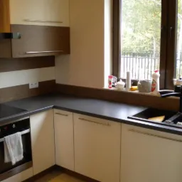 Nowoczesna kuchnia z białymi frontami szafek, ciemnym blatem i elementami drewna, okno z widokiem na zieleń, czarna bateria kuchenna i zlew.