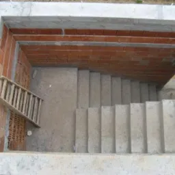 Betonowe schody w trakcie budowy, widoczne z góry, z drewnianą drabiną opartą o ceglaną ścianę, surowy stan wykończenia.