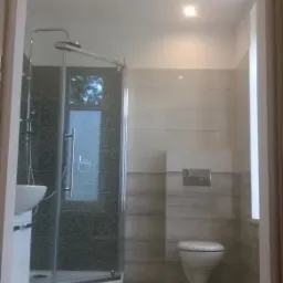 Wykończona łazienka z kabiną prysznicową narożną z czarną mozaiką na ścianach, umywalką na szafce, podwieszaną toaletą i beżowymi płytkami podłogowymi oraz ściennymi.