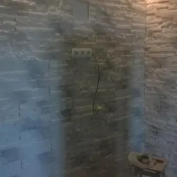 Ściana wyłożona dekoracyjnym kamieniem w odcieniach szarości i beżu, z widocznym gniazdkiem elektrycznym i otworem na instalację, w tle narzędzia i wiadro na taborecie.