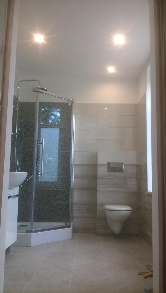 Wykończona łazienka z kabiną prysznicową narożną z czarną mozaiką na ścianach, umywalką na szafce, podwieszaną toaletą i beżowymi płytkami podłogowymi oraz ściennymi.