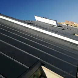 Montaż czarnej blachy na dachu, widoczny pracownik w niebieskim kasku i żółtej kamizelce odblaskowej, fragmenty konstrukcji i elementy mocujące.