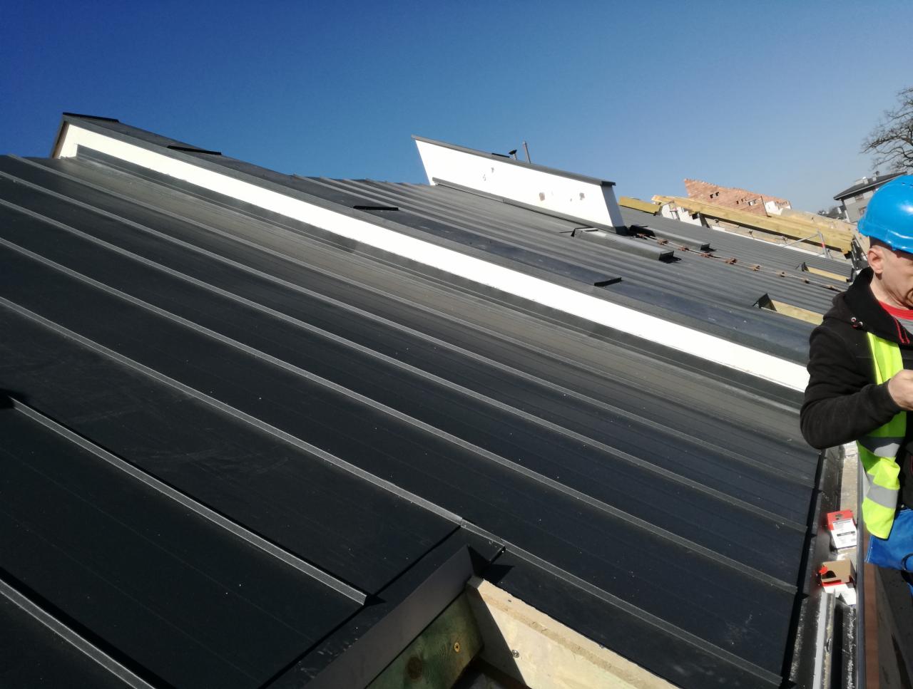 Montaż czarnej blachy na dachu, widoczny pracownik w niebieskim kasku i żółtej kamizelce odblaskowej, fragmenty konstrukcji i elementy mocujące.