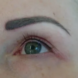 Zbliżenie na oko z precyzyjnie wykonanym permanentnym makijażem brwi i kreską na powiece, widoczne pojedyncze rzęsy i zielona tęczówka.