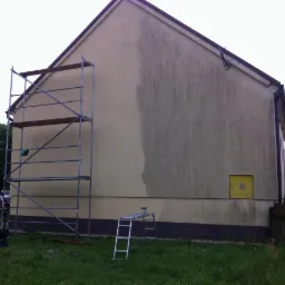 Boczna ściana domu, częściowo umyta, z widocznym rusztowaniem i drabiną, ukazująca kontrast między czystą a brudną powierzchnią.