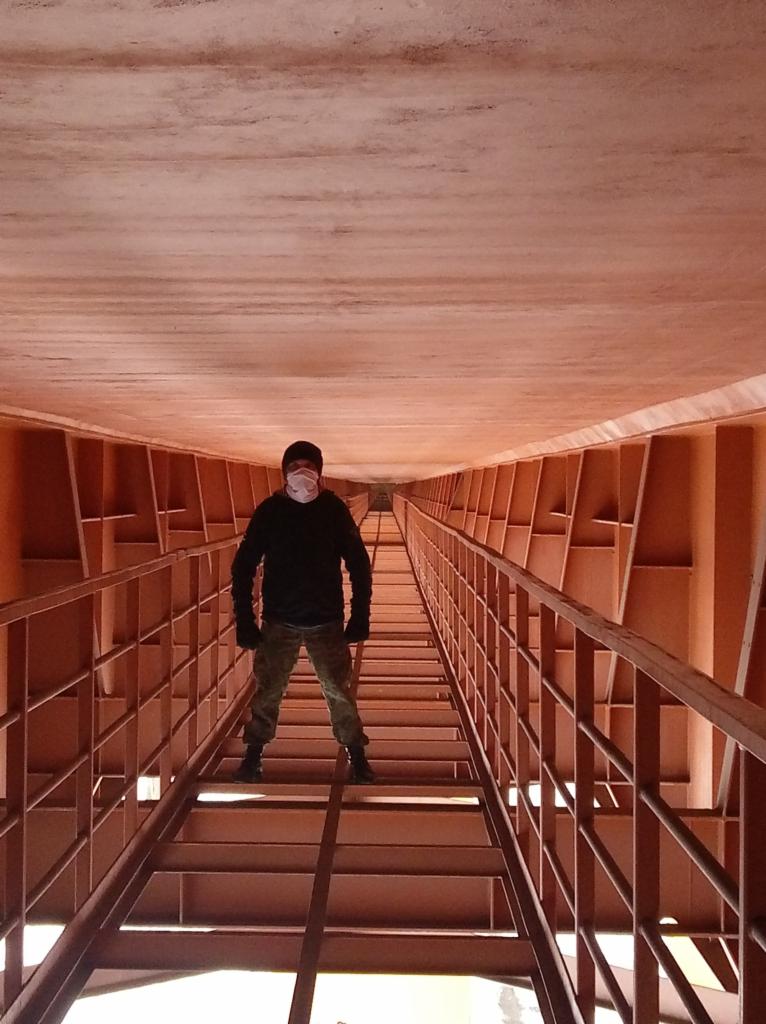 Osoba w masce na schodach konstrukcji stalowej, widok z dołu, perspektywa zbieżna, oświetlenie górne, elementy metalowe w kolorze ceglastym.