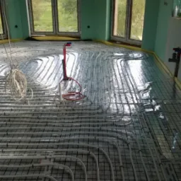 Rozłożona instalacja ogrzewania podłogowego na czarnej siatce w surowym wnętrzu z oknami wychodzącymi na zewnątrz.