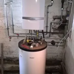 Biały piec gazowy Vaillant zawieszony na ścianie z widocznymi rurami, pod nim biały zasobnik ciepłej wody. Na zasobniku leżą złączki i narzędzia hydrauliczne.