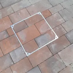 Ułożona kostka brukowa w odcieniach czerwieni i szarości, z wkomponowaną kwadratową studzienką rewizyjną z metalową ramą.