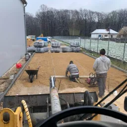 Prace przygotowawcze podłoża pod nawierzchnię: dwóch robotników wyrównuje piasek, jeden na kolanach, drugi z zagęszczarką, widok z kabiny maszyny budowlanej, w tle palety z kostką i budynek.