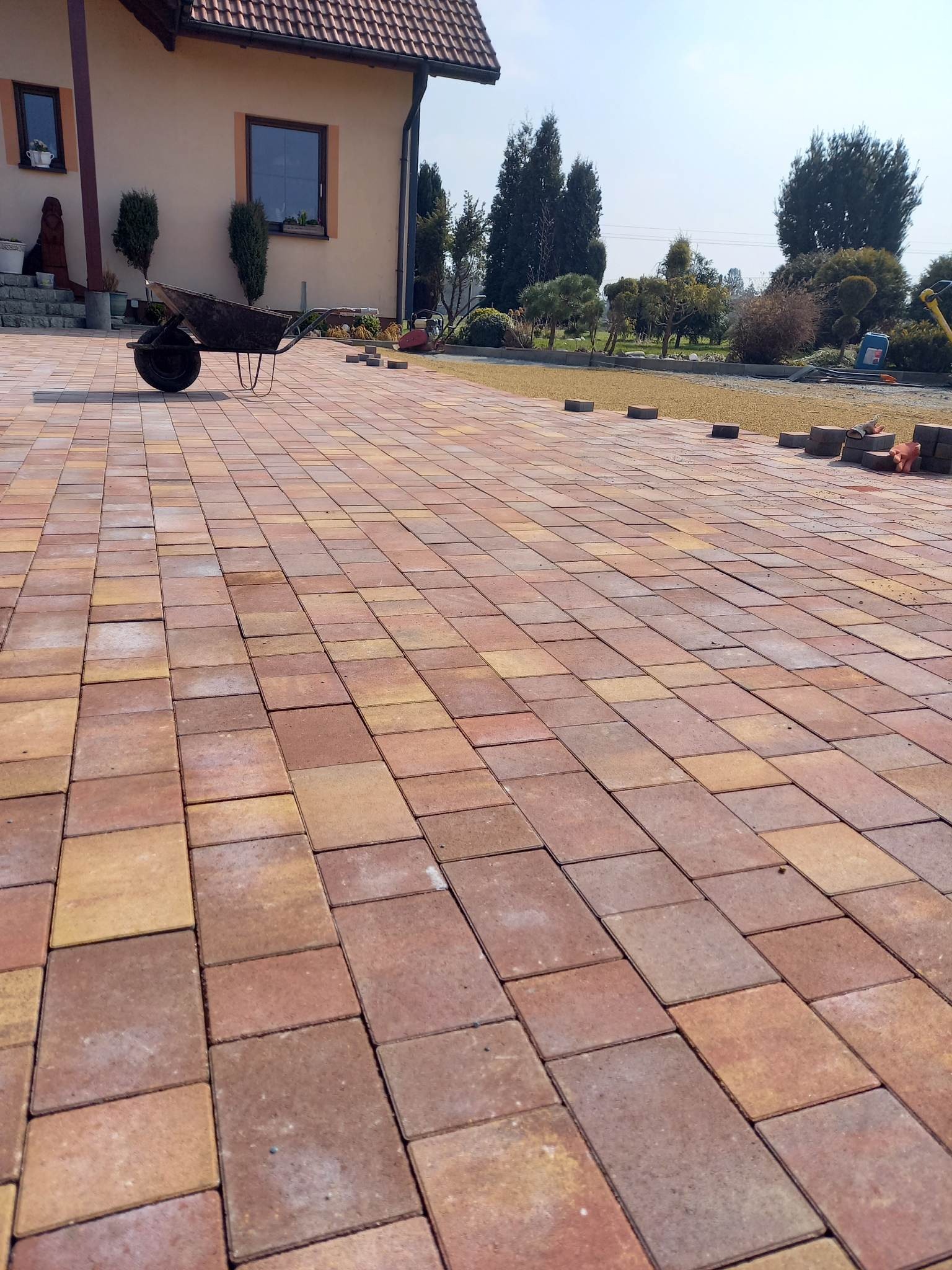 Nowo ułożona, kolorowa kostka brukowa na podjeździe, w tle dom jednorodzinny i taczka z narzędziami.