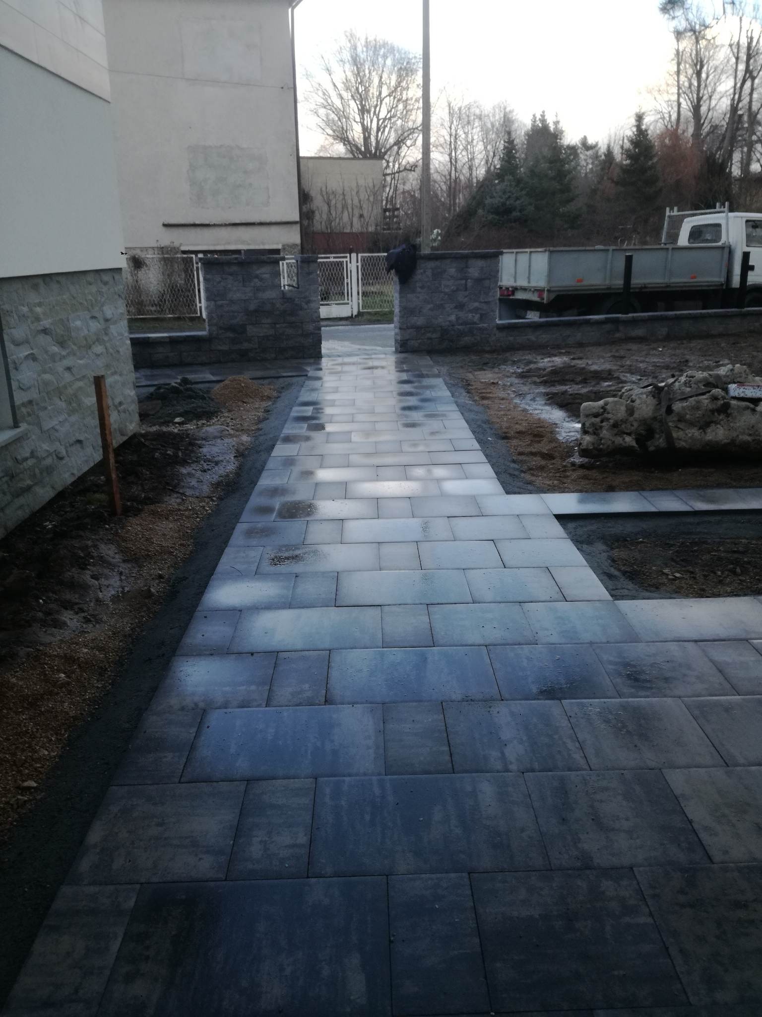 Świeżo ułożona, ciemnoszara kostka brukowa prowadząca do domu, z widocznymi fragmentami ogrodu i zaparkowaną ciężarówką w tle.