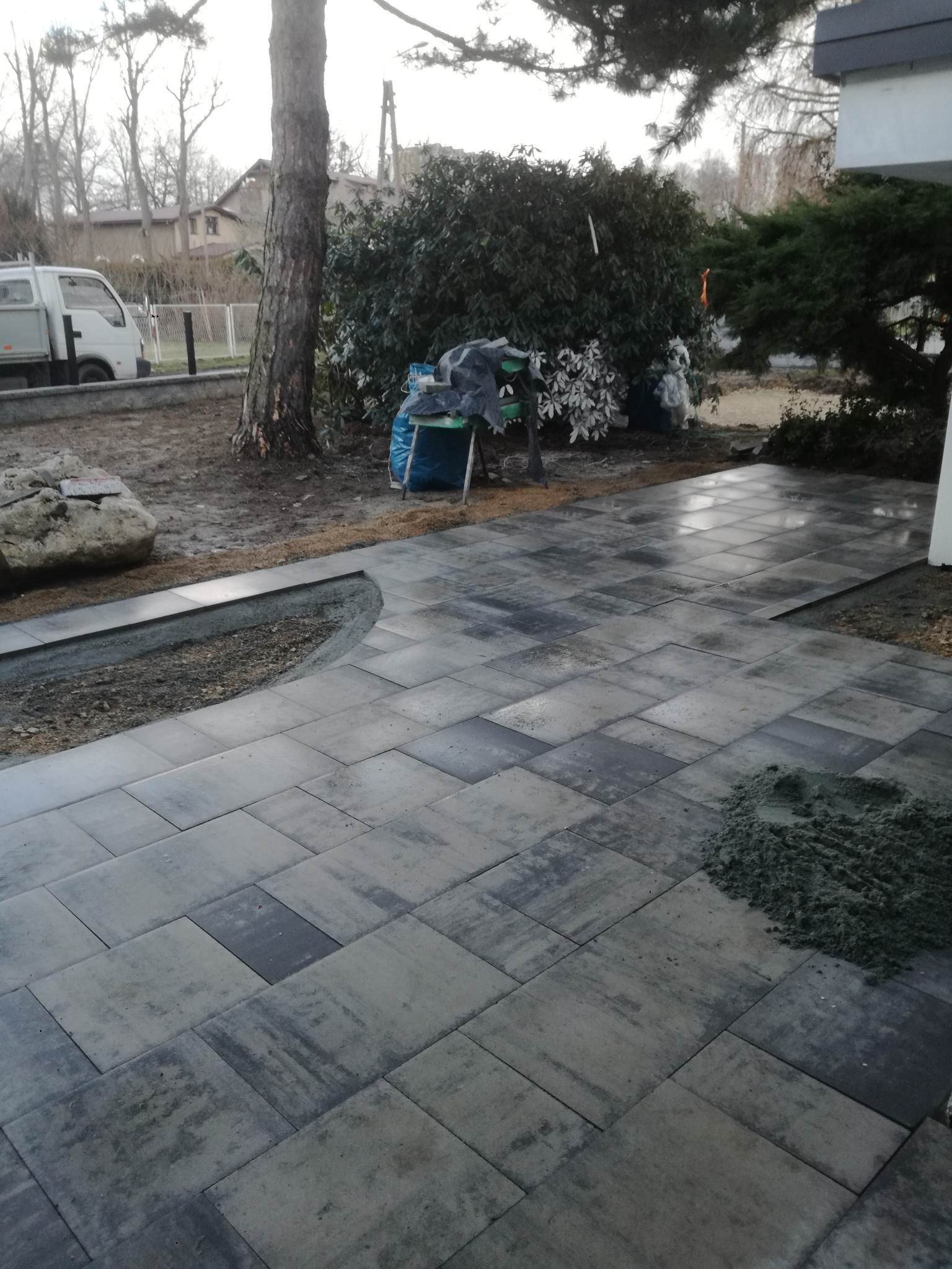 Świeżo ułożona, szara kostka brukowa na posesji, widoczny fragment rabaty, drzewo i samochód dostawczy w tle.