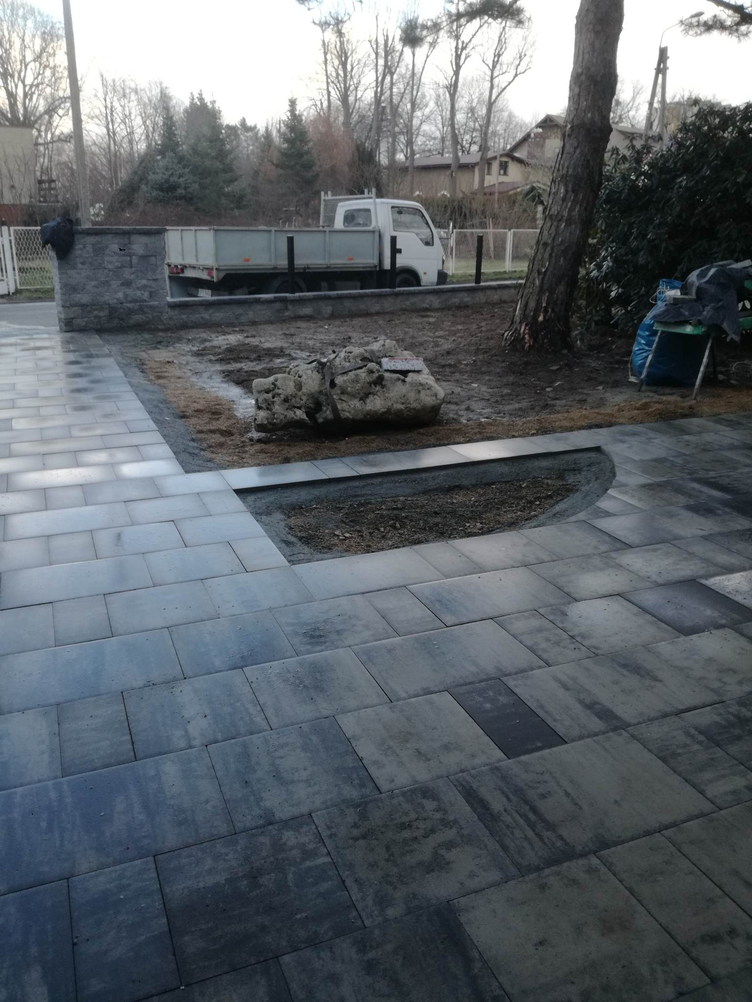 Nowo ułożona, szara kostka brukowa w trakcie prac wykończeniowych, z widocznym wycięciem pod element dekoracyjny, na tle ogrodzenia z kamienia i zaparkowanej ciężarówki.