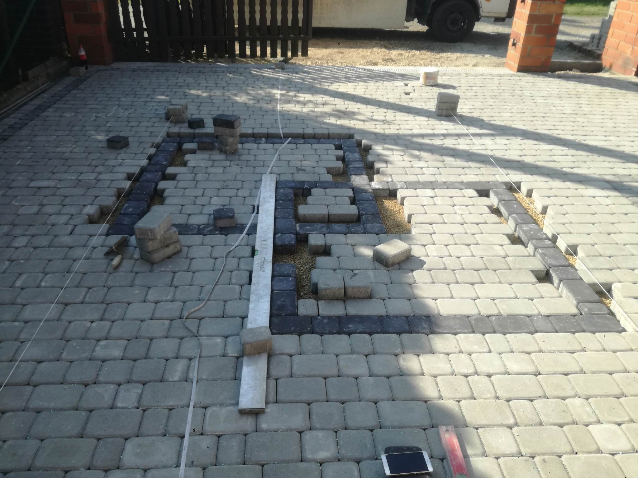 Układanie kostki brukowej na podjeździe, widoczne czarne i szare kostki, sznurek wyznaczający linię, poziomica i smartfon leżące na kostce brukowej, częściowo ułożona nawierzchnia.