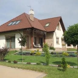 Ujęcie domu jednorodzinnego z brązowym dachem, zadbanym trawnikiem i ozdobnymi nasadzeniami krzewów oraz drzewek.