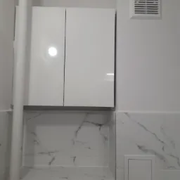 Biała szafka łazienkowa z dwoma drzwiczkami nad wnęką wyłożoną marmurowymi płytkami, widoczny pionowy biały rurociąg i kratka wentylacyjna na ścianie.